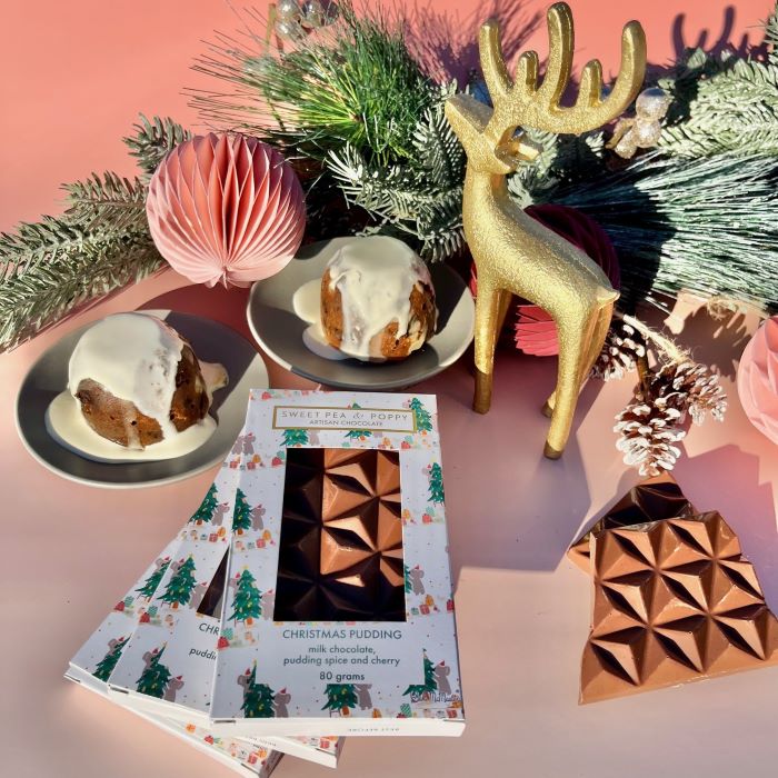 Christmas-themed items including a gold deer figurine, festive decorations, and a Christmas pudding artisan chocolate bar on a pink background.