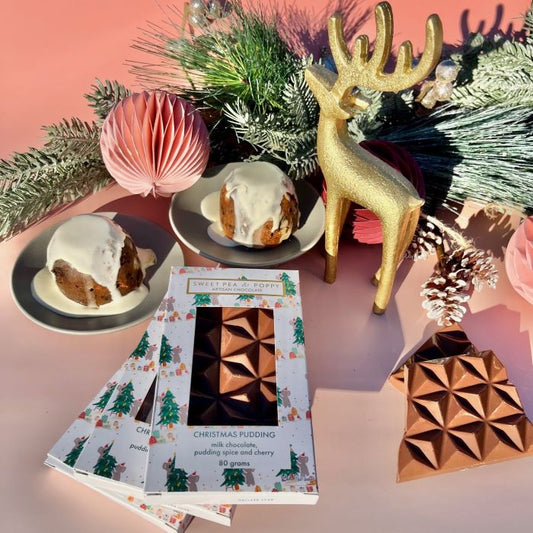 Christmas-themed items including a gold deer figurine, festive decorations, and a Christmas pudding artisan chocolate bar on a pink background.